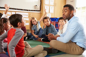 Black male teacher sitting on floor with circle of kids around him